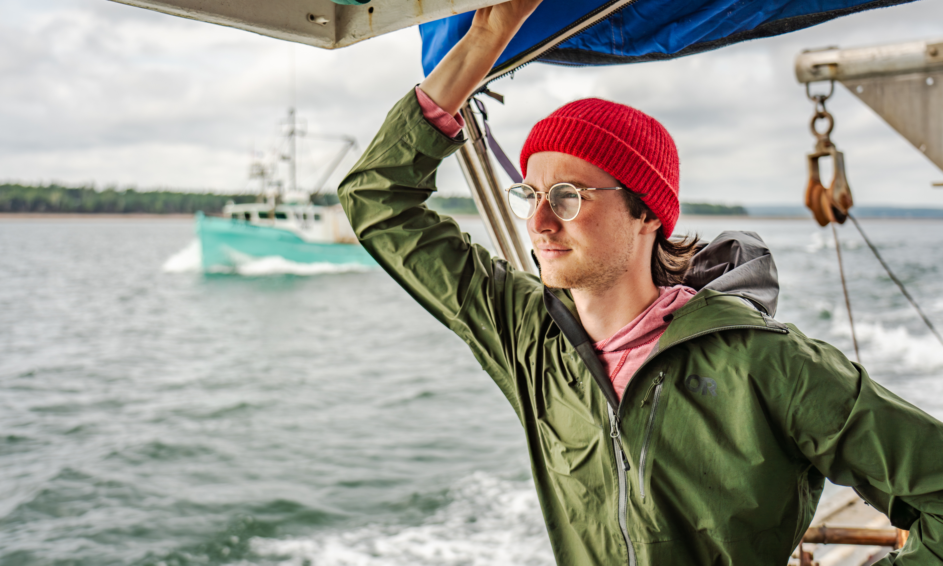 A student in a red beanie and green jacket stands on a boat deck, looking thoughtfully out at the sea with another vessel in the background. The scene captures a contemplative moment during a marine biology expedition.