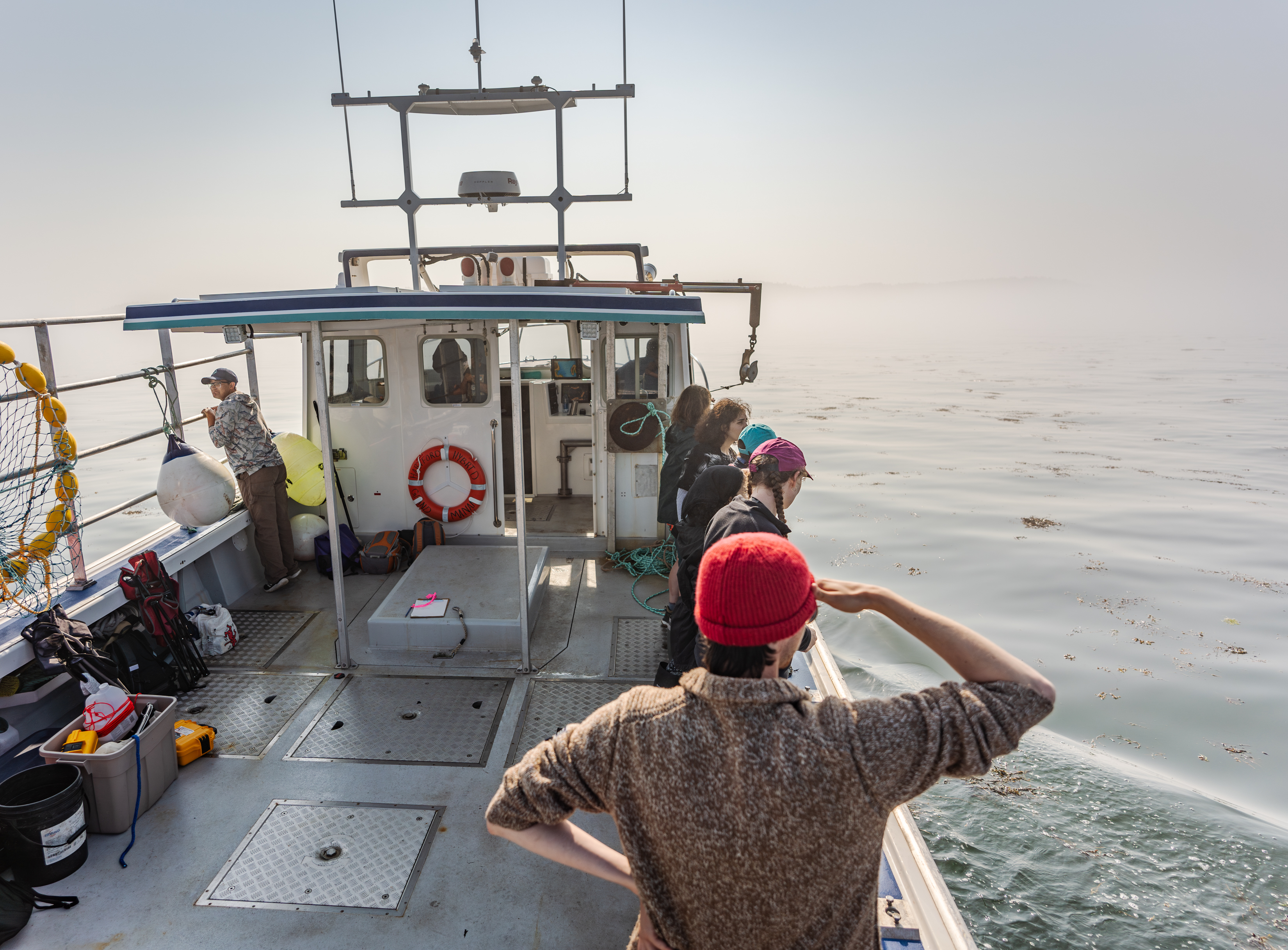 A group of students on the deck of a research vessel looking out over a misty ocean. Some students are pointing and observing the water as the boat sails forward.